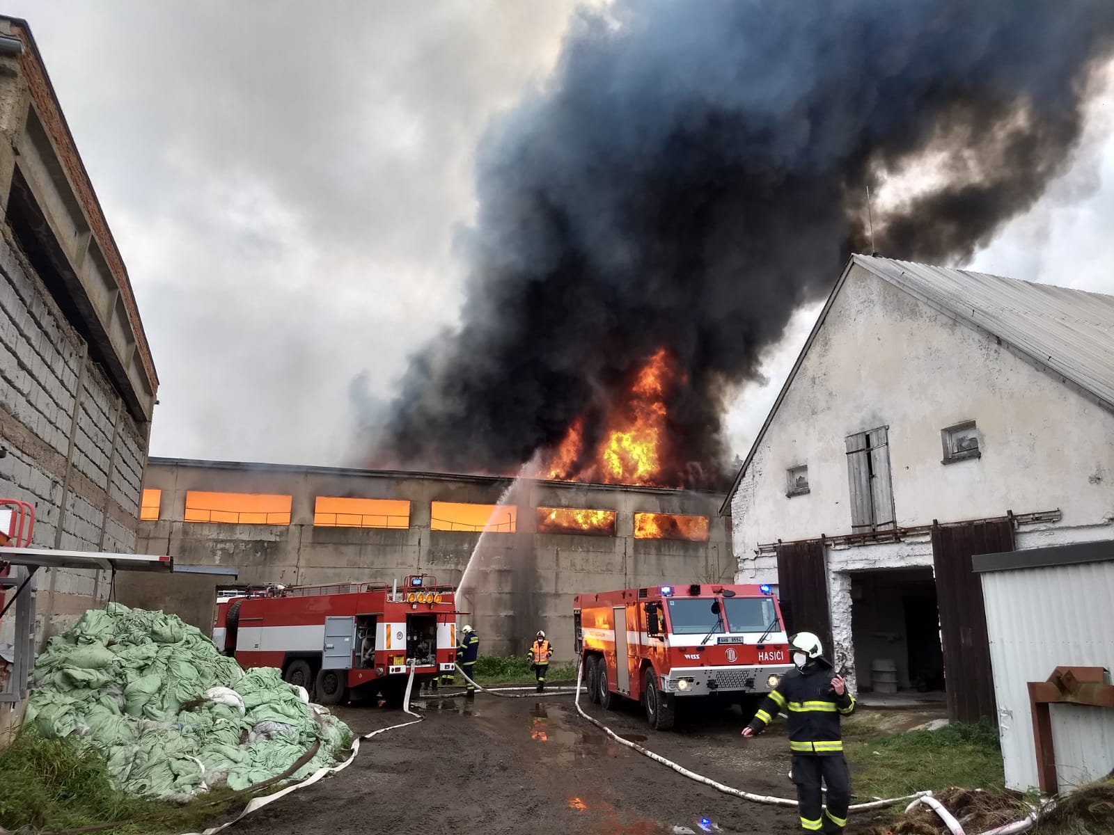 SČK_Požár haly s uskladněným senem v Bartošovicích_2 hasičské vozy stojí před hořící halou, ze které šlehají plameny a černý dým_4 hasiči v akci.jpg